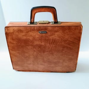May include: A vintage brown briefcase with a textured exterior. The briefcase has a black handle and a gold-coloured clasp. The brand name "Strandlite" is displayed on a small metal plate on the front.