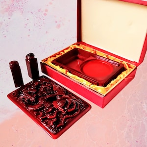May include: A red, intricately carved stamp set featuring a dragon motif. The set comprises a rectangular stamp, two smaller stamps, and a red ink pad, all housed in an open red box with a gold satin lining. The box is open, displaying the contents.