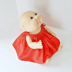 May include: A vintage doll with a bald head, blue eyes, and a red dress. The doll is sitting with its arms and legs extended. The red dress has a square neckline and ties at the shoulders. The doll's skin is a pale peach colour.