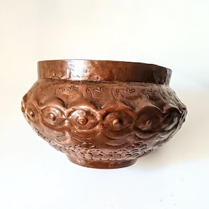 May include: A copper-toned, handmade bowl with detailed embossed patterns. The bowl displays repeating eye-like designs and floral motifs around the exterior. The vessel has a wide, open top and a rounded base, showcasing a rustic, antique aesthetic.