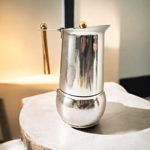 May include: A stainless steel stovetop espresso maker with a gold-coloured handle and top knob. The coffee pot has a bulbous base and a slender upper chamber. The handle is vertical and the top knob is a small sphere.