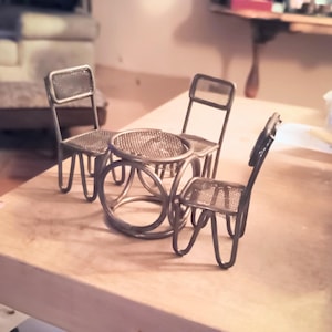 May include: A miniature metal patio set comprising a round table and three chairs. The table and chairs feature a mesh design on the seats and tabletop. The set is made of dark metal and rests on a light-coloured wooden surface.