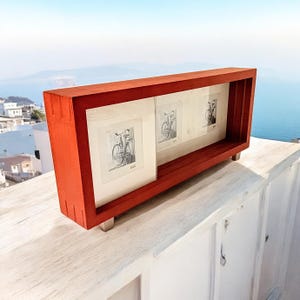 May include: A red wooden picture frame, rectangular in shape, with three sections, each holding a monochrome bicycle sketch. The frame is supported by small, silver feet. The background features a coastal scene with blue sea and sky.