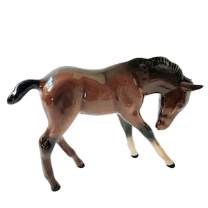 May include: A ceramic horse figurine in a poised stance. The horse is finished with a glossy glaze in shades of brown, black, and cream. The horse's head is bent down, and its tail is curved.