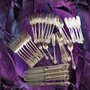 May include: A large collection of silver-toned cutlery including multiple forks, spoons, and knives. The set features ornate, decorative handles with intricate detailing. The pieces are arranged in a group, showcasing various sizes of dinner forks, salad forks, soup spoons, teaspoons, butter knives, and dinner knives.
