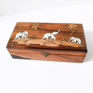 Peut inclure: Une boîte rectangulaire en bois avec un couvercle à charnière. La boîte est décorée d'une scène sculptée d'éléphants, de palmiers et d'oiseaux. Les éléphants sont blancs et la boîte a un fermoir en laiton.