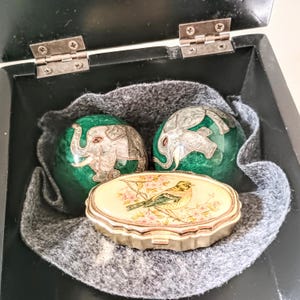 May include: A black box with a grey felt lining contains two green spheres decorated with elephants and a small, oval trinket box. The trinket box has a bird and floral design.