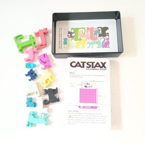 Peut inclure: Jeu de puzzle Cat Stax avec des pièces en forme de chat colorées. Le jeu comprend un plateau noir, des cartes défis et une carte blanche avec des instructions. Les pièces sont de différentes couleurs, dont le vert, le rose, le bleu, le jaune et le gris.