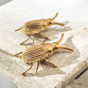 May include: Two gold-coloured beetle figurines. Each beetle has a segmented body, six legs, and curved antennae. The beetles are displayed on a light-coloured, textured surface. The figurines are likely decorative items.