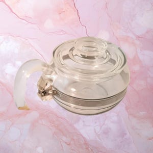 May include: A clear glass teapot featuring a rounded body, a matching glass lid with a central knob, and a curved glass handle attached with a silver-toned metal band. The design includes a horizontal silver-toned stripe around the centre.