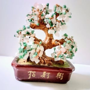 May include: A decorative bonsai tree sculpture with a brown trunk and branches, adorned with green, white, and pink gemstone leaves. The tree sits in a rectangular, dark red pot with gold Chinese characters.