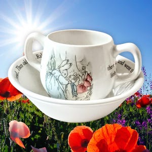 May include: A white ceramic cup and saucer set, decorated with a Peter Rabbit illustration. The cup has two handles and the saucer is bowl-shaped. The set features a rabbit scene and text, set against a floral background.