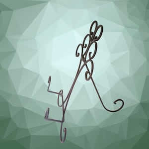 May include: A dark brown metal display stand featuring a tall, arched back with intricate scrollwork patterns and two front-facing hooks designed to hold items securely in an upright position, supported by a hinged rear leg for stability.