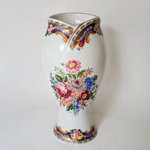 May include: A white ceramic jug featuring a central floral bouquet with pink, red, and blue flowers. The rim and base are decorated with ornate gold, blue, and burgundy scrollwork patterns. The jug has a smooth, glossy finish throughout.