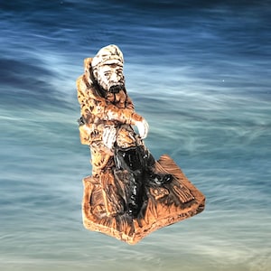 May include: A small figurine of a bearded sailor wearing a cap and heavy coat, seated on a textured base. The figure is rendered in shades of brown and tan with black boots, capturing a weathered appearance with detailed facial features and clothing.