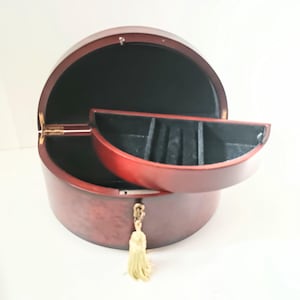 Peut inclure: Un écrin à bijoux rond en bois, de couleur brun rougeâtre. Le coffret est ouvert, révélant un intérieur noir et un petit plateau semi-circulaire avec des compartiments. Une gland doré est suspendu à l'avant.