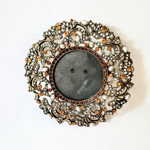 May include: A circular metal picture frame with an antique bronze finish, featuring an intricate openwork scroll design. The frame is embellished with clear white crystals, small orange gemstones, and white faux pearls throughout the border.