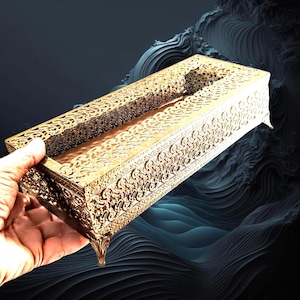 May include: A rectangular gold-coloured metal tissue box cover featuring an intricate, laser-cut filigree pattern across all four sides and the top surface. The box is supported by four small, ornate, curved feet at each corner of the base.