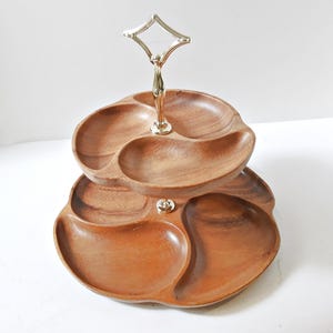 May include: A two-tiered serving tray with a gold-coloured handle. The trays are made of wood and have a three-sectioned design. The wood has a warm, brown tone, and the handle has a decorative, geometric top.
