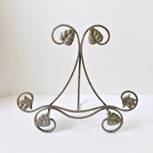 May include: A decorative metal display stand with a bronze finish. The stand features a curved design with leaf accents and a three-legged base. The top has two curved supports with leaf details. The stand is designed to hold a plate or picture.