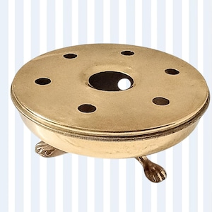 May include: A gold-coloured, round incense burner with small holes on the top. It has a central opening and is supported by three decorative feet. The design is simple and elegant. The burner is likely made of brass or a similar metal.
