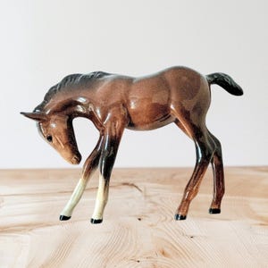 May include: A ceramic foal figurine with a glossy finish. The foal is depicted in a poised stance, with a brown body, black mane and tail, and legs that are white and black. The background is neutral.