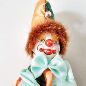Peut inclure: Poupée clown vintage avec un visage en porcelaine peint, un nez rouge et des lèvres rouges. La poupée a des cheveux en fausse fourrure brune, des yeux bleus et un nœud papillon bleu clair. Elle porte un chapeau coloré avec une boule argentée.