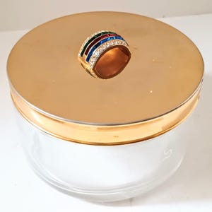 May include: A clear glass jar with a gold-coloured lid. The lid is topped with a gold-toned ring adorned with red, green, blue, and clear gemstones. The jar has a gold rim.