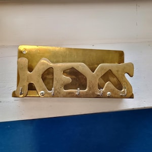 May include: A brass key holder with the word "KEYS" cut out. The key holder has a rectangular backplate and five hooks below the letters. The key holder is mounted on a white and blue surface.