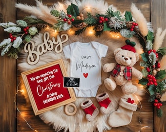 Könnte beinhalten: Weihnachtliche Babyankündigung mit einem weißen Body mit dem Namen "Madison" und einem roten Herz. Ein Teddybär, Babysocken und eine gerahmte Ankündigung sind ebenfalls enthalten. Die Ankündigung lautet: "Wir erwarten das beste Geschenk dieses Weihnachten, Ankunft Dezember 2025".
