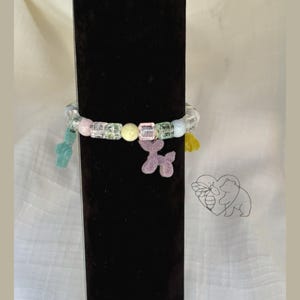 May include: A beaded bracelet with clear, pink, green, and yellow beads. The bracelet has a clear elastic band and features charms of a green gummy bear, a purple balloon dog, and a yellow tassel. The bracelet is displayed on a black stand.