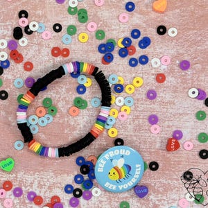 May include: A black beaded bracelet with rainbow-colored accents and a round button with a bee and the text "BEE PROUD BEE YOURSELF". Colorful beads and heart-shaped embellishments are scattered around the bracelet and button.