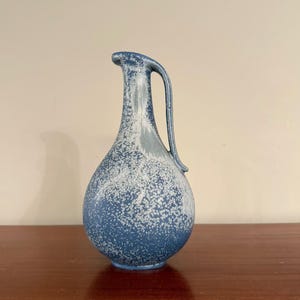 Może przedstawiać: Ceramiczny dzbanek z pękatą podstawą i smukłą szyjką, pokryty niebieskim szkliwem z białym nakrapianym wzorem. Naczynie posiada pojedyncze, zakrzywione ucho biegnące od krawędzi do ramienia, co nadaje mu charakterystyczną sylwetkę i fakturę.