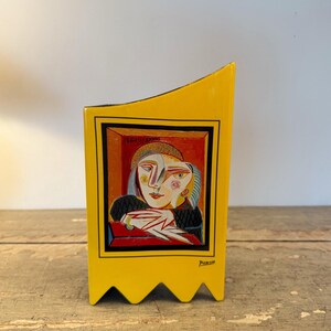 Può includere: Un vaso di ceramica giallo con un bordo nero e un dipinto in stile Picasso di due figure. Il dipinto è incorniciato in un rettangolo rosso. Il vaso ha un bordo inferiore a zig-zag e la firma dell'artista.
