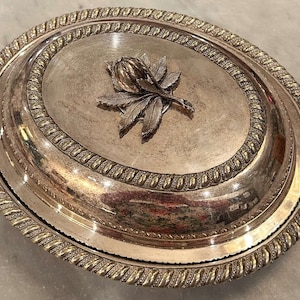May include: Silver oval serving dish with a raised floral design on the lid. The dish has a gold rim with a rope-like pattern.