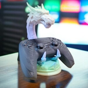 May include: A 3D printed game controller stand featuring a dragon head design. The dragon is light purple and green, and the controller stand is black. The stand has a base that is light green and white.