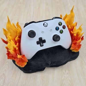 May include: A white video game controller with black buttons and a black base, surrounded by orange and yellow flames. The controller is resting on a black stand.