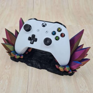 May include: A white Xbox controller sits in a 3D printed stand. The stand is black with a textured surface and features colourful, iridescent crystals on either side.