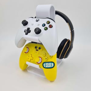 May include: White stand with two video game controllers and a black headset. The top controller is white and the bottom controller is yellow with a smiley face and the text "NANA NANA".