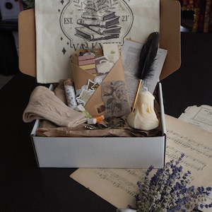 May include: A white box filled with book club themed items. Includes a tote bag with text, a feather pen, a candle, stickers, a pair of scissors, a pair of socks, and a bundle of lavender tied with a silver ribbon.