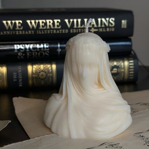 May include: Ivory-colored candle shaped like a veiled bust, with a wick at the top. The candle sits on sheet music, with a stack of black books behind it. The books have gold lettering that reads "WE WERE VILLAINS" and "PSYCHE & EROS".