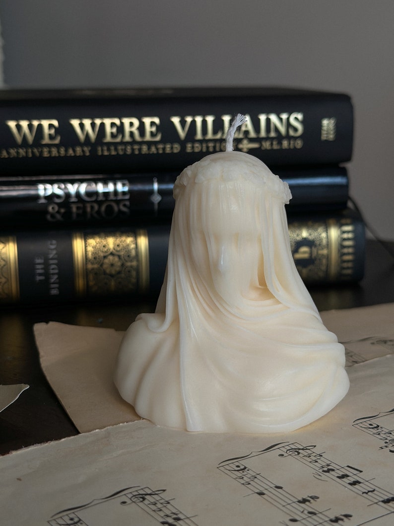 May include: Ivory-colored candle shaped like a veiled bust, with a wick at the top. The candle sits on sheet music, with a stack of black books behind it. The books have gold lettering that reads "WE WERE VILLAINS" and "PSYCHE & EROS".