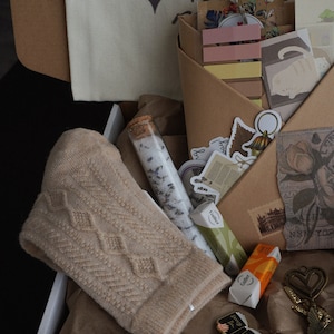 May include: A gift box filled with a beige knit hat, bath salts, stickers, and stationery. The box contains a variety of items, including a small pin, a key-shaped charm, and wrapped candies. A canvas tote bag with the word "VALKYRIE" is visible.