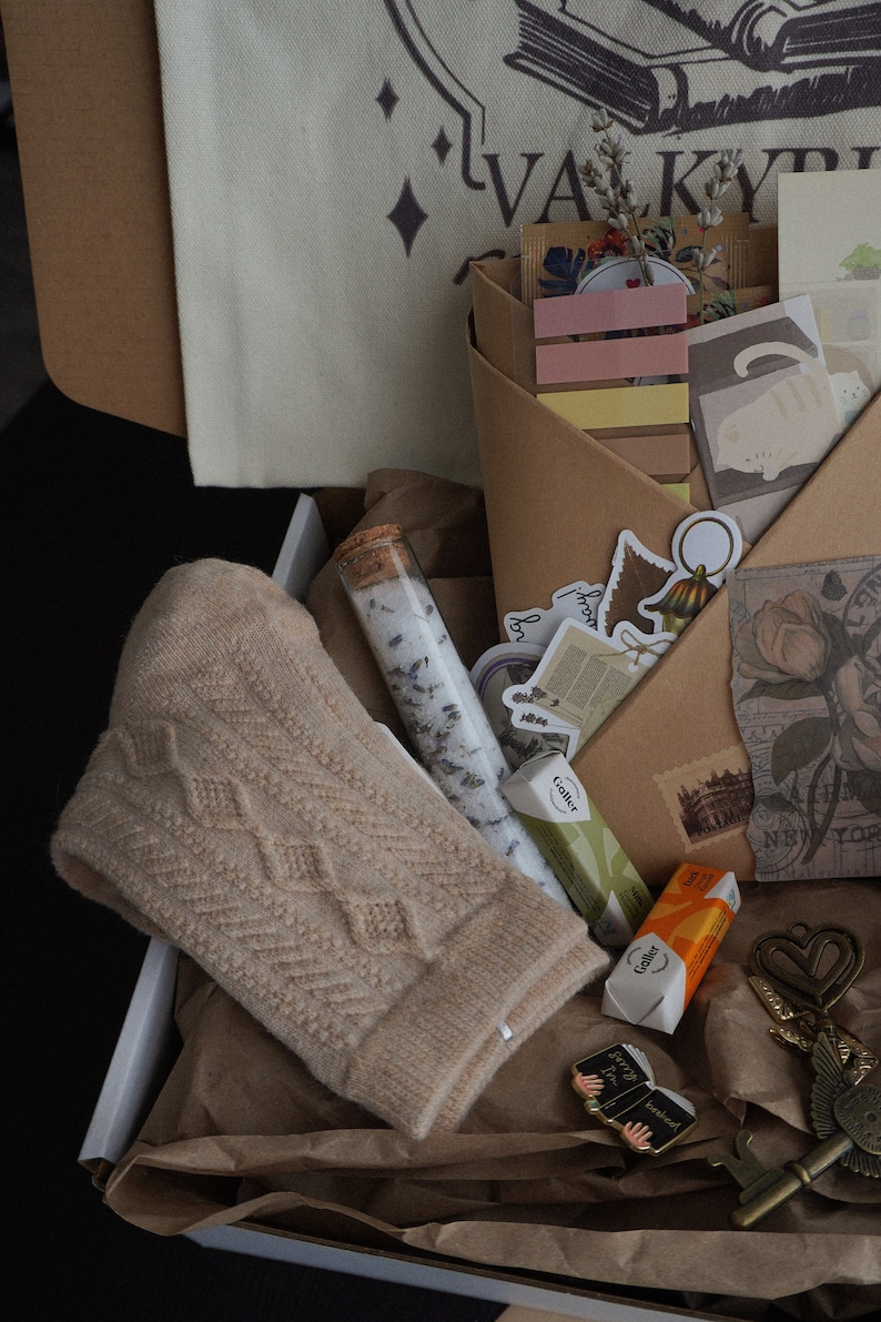 May include: A gift box filled with a beige knit hat, bath salts, stickers, and stationery. The box contains a variety of items, including a small pin, a key-shaped charm, and wrapped candies. A canvas tote bag with the word "VALKYRIE" is visible.