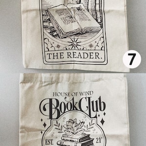 May include: Two beige canvas tote bags with black graphic designs. One features an open book and the text "THE READER." The other says "HOUSE OF WIND Book Club" and "VALKYRIE reading room."