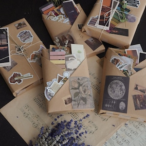 May include: Several packages wrapped in brown paper, adorned with stickers and tied with twine. The packages contain various ephemera, including postcards, stickers, and small prints. A bouquet of lavender and sheet music are also present.