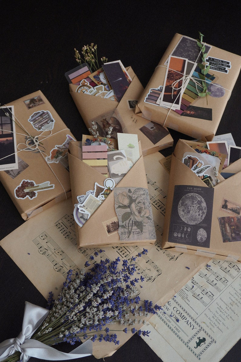 May include: Several packages wrapped in brown paper, adorned with stickers and tied with twine. The packages contain various ephemera, including postcards, stickers, and small prints. A bouquet of lavender and sheet music are also present.