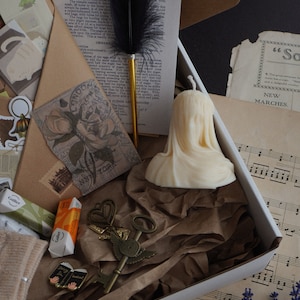 May include: A white box filled with vintage-themed items. Includes a cream-colored candle, a black feather quill, antique keys, old paper ephemera, and small wrapped candies. The box is lined with brown paper.