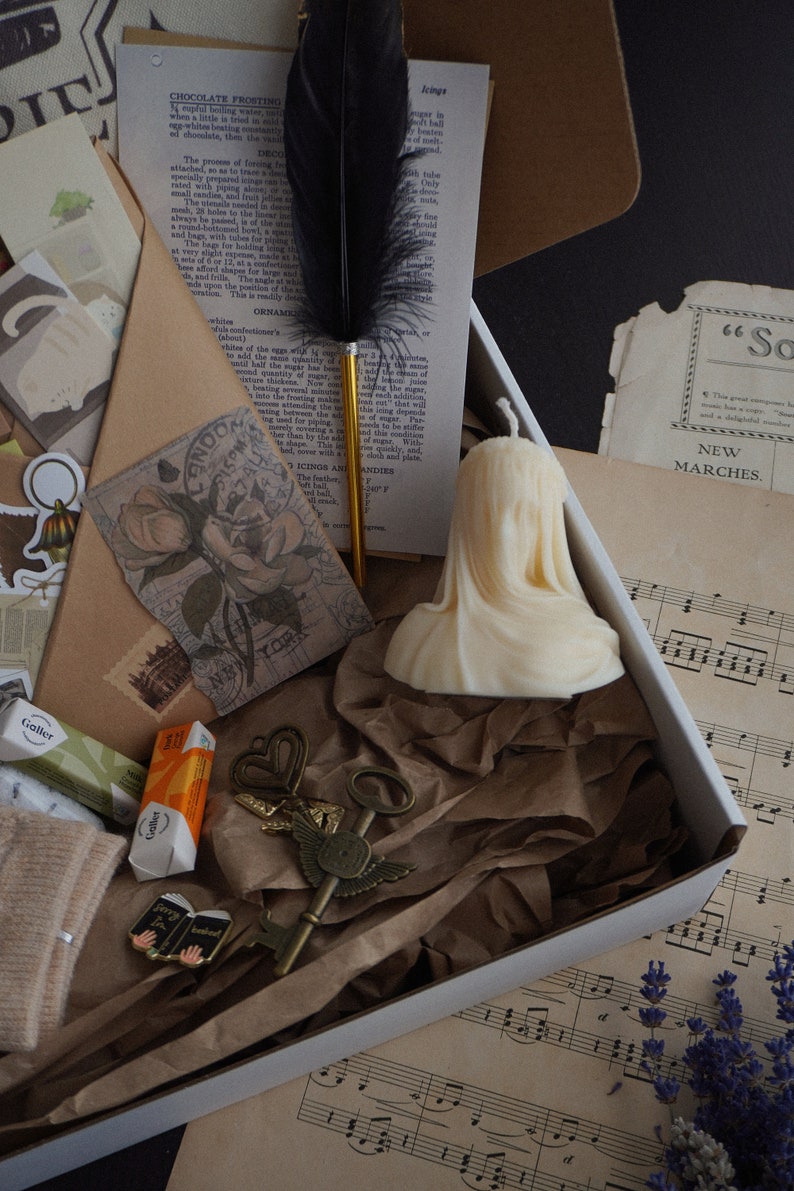 May include: A white box filled with vintage-themed items. Includes a cream-colored candle, a black feather quill, antique keys, old paper ephemera, and small wrapped candies. The box is lined with brown paper.