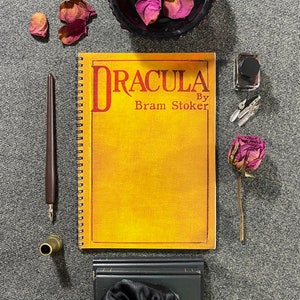 Könnte beinhalten: Ein Spiralnotizbuch mit dem Titel "DRACULA by Bram Stoker" in roter Schrift auf einem gelben Einband. Das Bild zeigt einen braunen Stift, ein Tintenfass, Federn und eine getrocknete Rose.